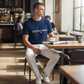 Hombre con camiseta azul marino U.S. POLO ASSN. de algodón y pantalones blancos sentado en una cafetería