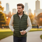 Hombre con chaleco acolchado verde oliva de www.treshop.com.co sobre suéter beige en un parque urbano al atardecer.