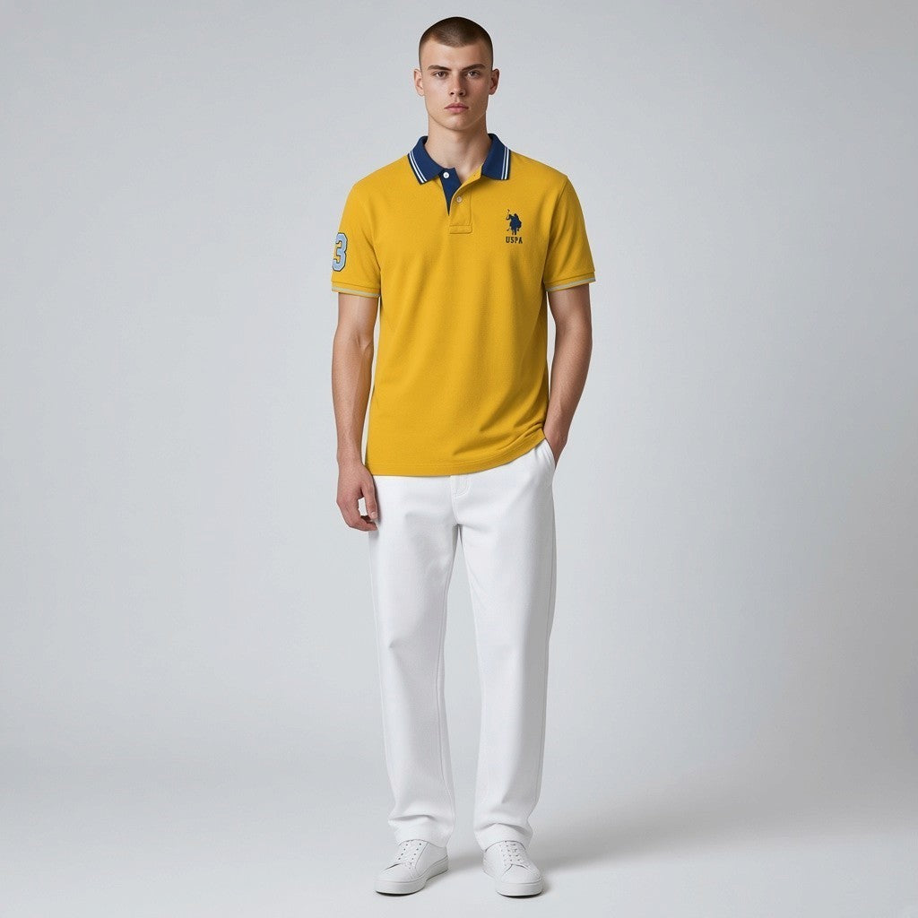 Hombre vistiendo polo amarillo mostaza www.treshop.com.co con cuello azul marino, logo bordado y pantalón blanco