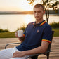Hombre con polo azul marino www.treshop.com.co con detalles en cuello y mangas, sentado al atardecer junto al lago.