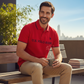 Hombre sonriente vistiendo polo rojo U.S. POLO ASSN. con detalles en contraste, sentado en un banco de madera.